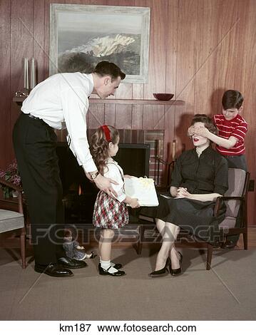 Stock Photo - 1950s FAMILY IN LIVING ROOM SURPRISING MOTHER WITH GIFT FATHER DAUGHTER HOLD GIFT SON COVERS MOMS EYES. Fotosearch
