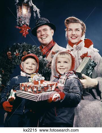 Stock Photography - 1950s FAMILY MAN WOMAN TWO KIDS HOLDING CHRISTMAS PRESENTS SMILING STANDING BENEATH LANTERN. Fotosearch