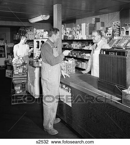 Stock Image - 1950S Man Farmer At Rural General Mercantile Store Counter Talking To Clerk Store Keeper At Cash Register. Fotosearch