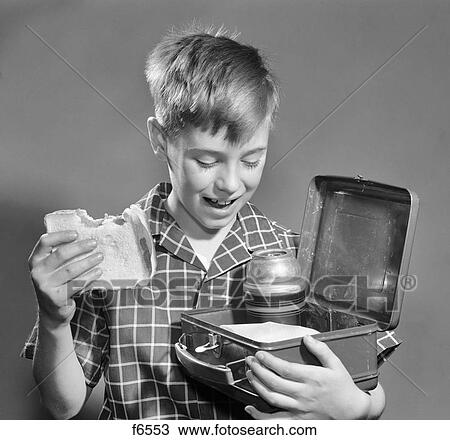 Stock Image - 1950s SMILING BOY EATING SANDWICH LOOKING INTO LUNCH BOX WITH THERMOS. Fotosearch