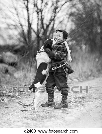 Stock Image - 1950s SMILING BOY WEARING WINTER COAT HAT SCARF BOOTS ICE SKATES SLUNG OVER SHOULDER HUGGING ENGLISH SPRINGER SPANIEL DOG. Fotosearch