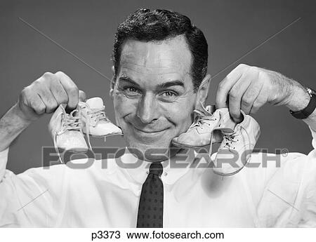 Stock Image - 1950S Smiling Portrait Man Holding One Pair Of Baby Booties In Each Hand Next To Face. Fotosearch