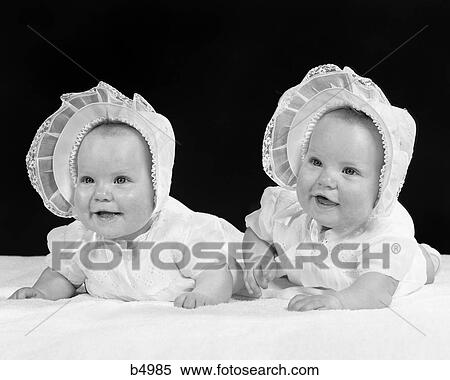 Stock Photography - 1950s TWIN BABY GIRLS WEARING BONNETS LYING ON THEIR STOMACHS SMILING. Fotosearch