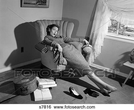 Stock Photograph - 1950s WOMAN EXHAUSTED FROM SHOPPING SLUMPING IN LIVING ROOM CHAIR SHOES OFF PACKAGES EVERYWHERE LOOKING AT CAMERA. Fotosearch