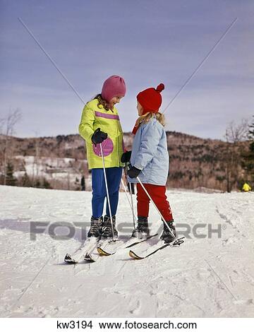 Picture - 1960s 1970s 2 JUVENILE GIRLS CHILDREN SKIING WEARING PASTEL PARKAS STANDING TALKING SISTERS. Fotosearch