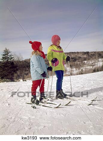 Stock Image - 1960s 1970s 2 JUVENILE GIRLS ON SKIS WEARING PASTEL PARKAS LOOKING UP SLOPE. Fotosearch