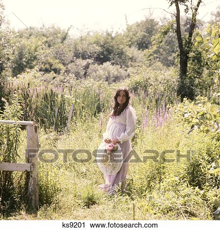 Stock Image - 1960S 1970S Pregnant Woman Standing In Field Of Flowers Granny Dress Big Hat. Fotosearch