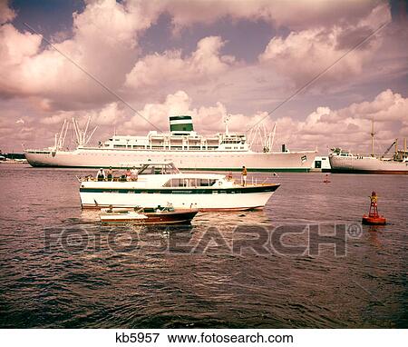 Stock Photo - 1960s 1970s SEVERAL FISHING AND PLEASURE MOTOR BOATS PASSING BY SMALL OCEAN GOING CRUISE SHIP MOORED IN HARBOR MARINA. Fotosearch