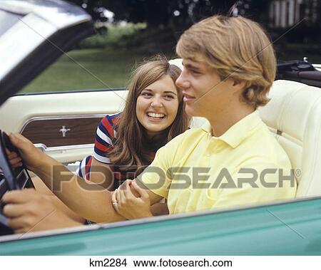Picture - 1960s 1970s SMILING TEENAGE COUPLE GIRL FLIRTING LOOKING AT HOLDING ARM OF BOY DRIVING CONVERTIBLE CAR. Fotosearch