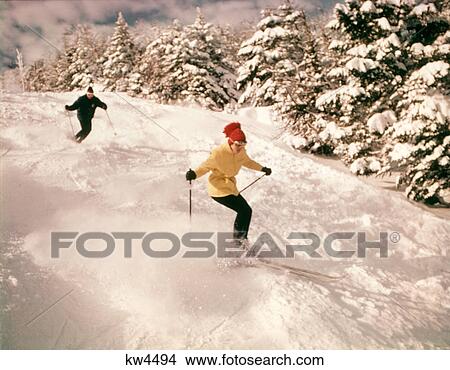 1960s 1970s TWO PEOPLE MAN WOMAN SKIING DOWN SNOWY SLOPE PINE TREES WOMAN WEARING YELLOW JACKET AND RED CAP View Large Photo Image Picture - 1960s 1970s TWO PEOPLE MAN WOMAN SKIING DOWN SNOWY SLOPE PINE TREES WOMAN WEARING YELLOW JACKET AND RED CAP. Fotosearch
