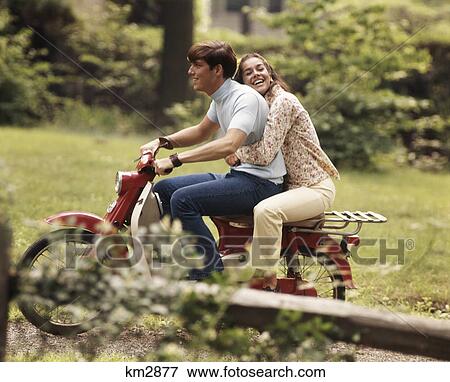 Stock Photo - 1960s 1970s YOUNG TEENAGE COUPLE RIDING ON MOTORBIKE BOY DRIVING GIRL HOLDING ON BEHIND LAUGHING. Fotosearch