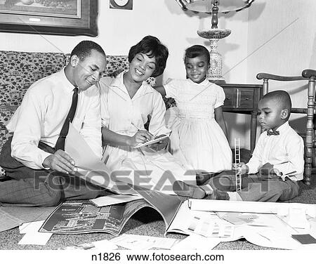 Stock Photograph - 1960s AFRICAN AMERICAN FAMILY LOOKING AT BLUEPRINTS PLANNING FOR A NEW HOME BOY GIRL MOTHER FATHER. Fotosearch