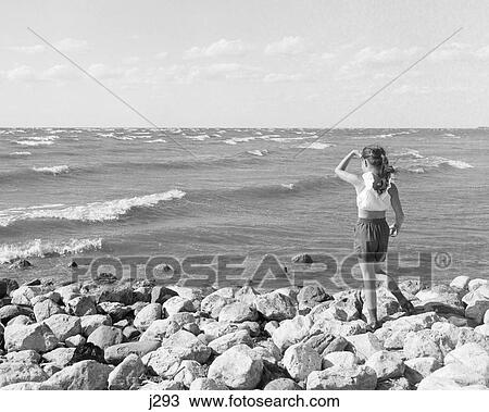 Stock Image - 1960s BACK VIEW OF LITTLE GIRL STANDING ON ROCKS LOOKING OUT TO HORIZON OF LAKE MICHIGAN WITH HAND SHADING EYES. Fotosearch