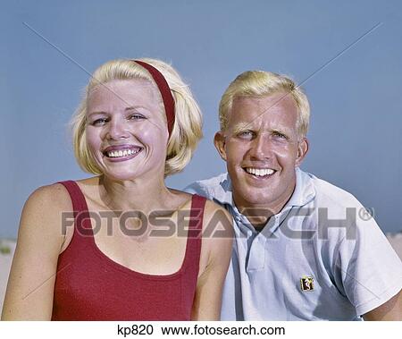 Stock Image - 1960s BLOND COUPLE AT SEASHORE BEACH SMILING LOOKING AT CAMERA. Fotosearch