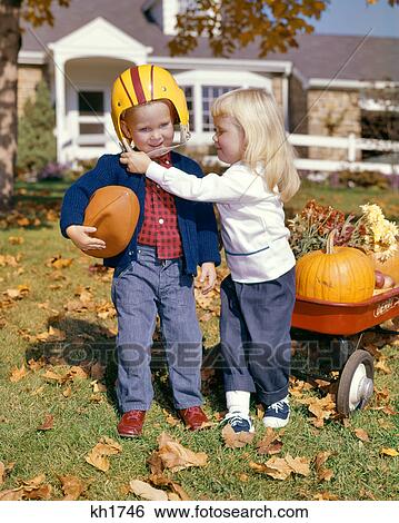 1960s BLONDE GIRL WEARING SADDLE OXFORDS HELPING BOY HOLDING FOOTBALL PUT ON HELMET AUTUMN YARD RED WAGON View Large Photo Image Stock Photograph - 1960s BLONDE GIRL WEARING SADDLE OXFORDS HELPING BOY HOLDING FOOTBALL PUT ON HELMET AUTUMN YARD RED WAGON. Fotosearch