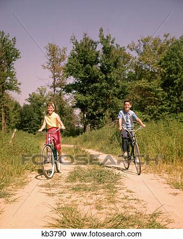 Stock Image - 1960s BOY AND GIRL RIDING BIKES ON COUNTRY LANE IN SUMMERTIME LOOKING AT CAMERA. Fotosearch