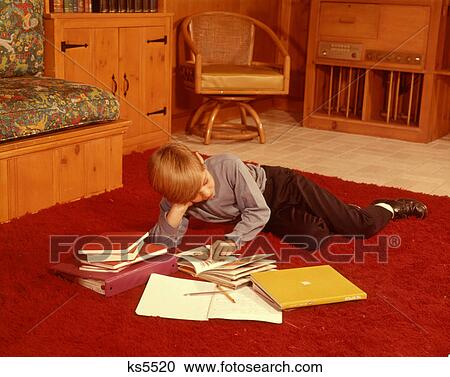 Stock Image - 1960s BOY READING STUDYING HOMEWORK LYING ON RED SHAG CARPET IN LIVING ROOM. Fotosearch