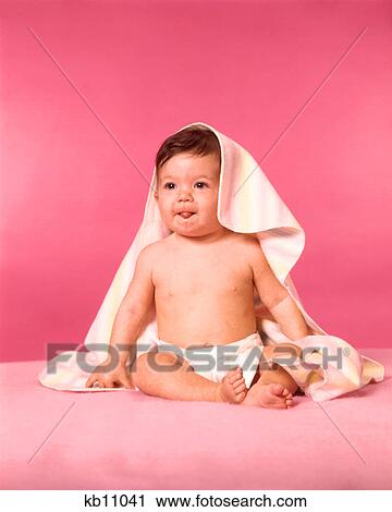Stock Image - 1960s BRUNETTE BABY WEARING DIAPER SITTING WITH BLANKET OVER HEAD STICKING OUT TONGUE. Fotosearch