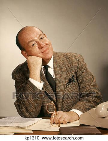 Stock Image - 1960s BUSINESSMAN AT OFFICE DESK HAND TO FACE HEAD TILTED LEANING ON FIST LOOKING AT CAMERA. Fotosearch