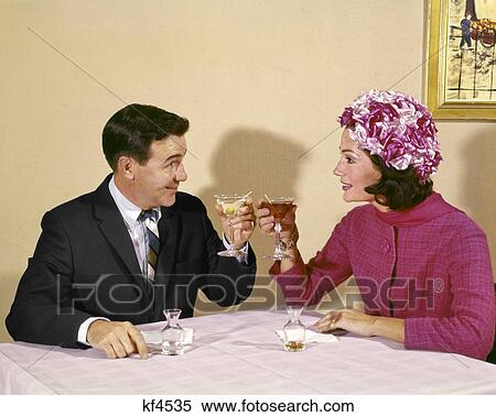 Stock Photography - 1960s COUPLE TOASTING EACH OTHER WITH WINE IN RESTAURANT WOMAN WEARING FLOWERED HAT. Fotosearch