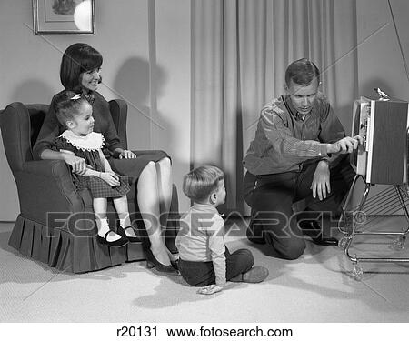 Stock Image - 1960s FAMILY OF FOUR WATCHING PORTABLE TELEVISION MOTHER DAUGHTER IN CHAIR FATHER TURNING TV CHANNEL DIAL SON SITTING ON RUG. Fotosearch