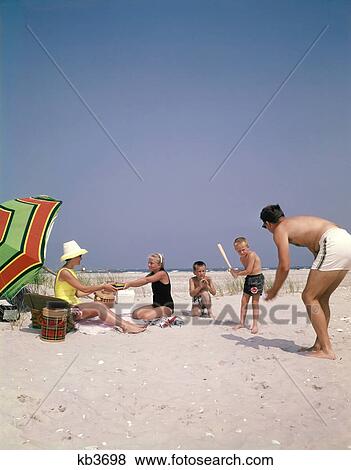 Stock Photo - 1960s FAMILY ON SUMMER SANDY BEACH MOTHER PICNICKING WITH DAUGHTER FATHER PLAYING PITCHING BASEBALL TO SONS. Fotosearch