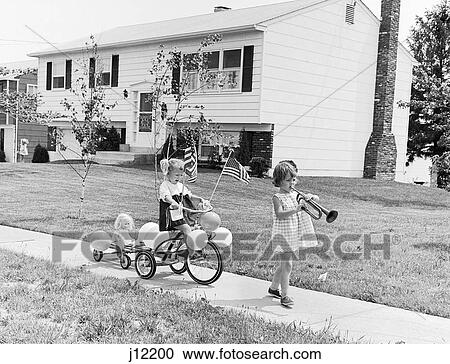 Stock Image - 1960s GIRL WITH BUGLE LEADING SISTER ON TRICYCLE DECORATED WITH BALLOONS AMERICAN FLAGS FOR JULY 4TH HOLIDAY SIDEWALK PARADE. Fotosearch
