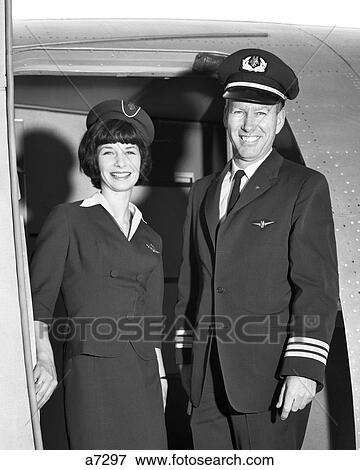 Stock Photo - 1960s MAN CO-PILOT AND WOMAN FLIGHT ATTENDANT STANDING SMILING IN DOOR OF AIRPLANE LOOKING AT CAMERA. Fotosearch