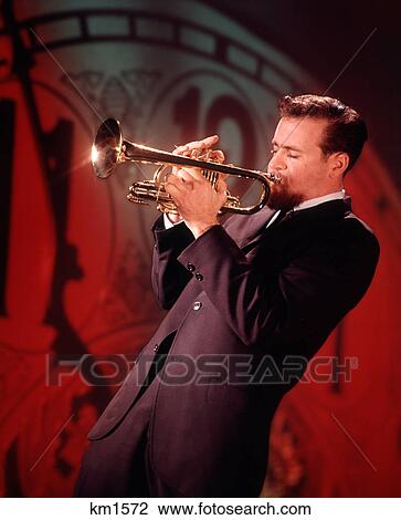 Stock Image - 1960s MAN JAZZ MUSICIAN PLAYING TRUMPET NEAR MIDNIGHT NEW YEARS CLOCK FACE IN BACKGROUND. Fotosearch