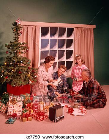 Stock Image - 1960s MAN WOMAN MOTHER FATHER TWO TEENAGERS BOY GIRL SON DAUGHTER AROUND CHRISTMAS TREE OPENING PRESENTS. Fotosearch