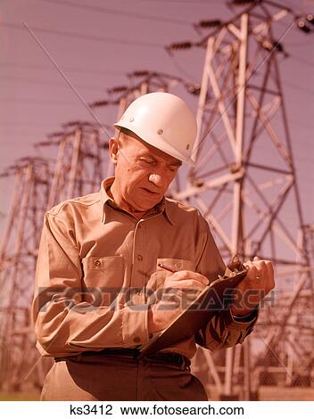 Stock Image - 1960s MAN WORKER CLIPBOARD HARD HAT OUTDOORS BY ELECTRICAL POWER LINES. Fotosearch