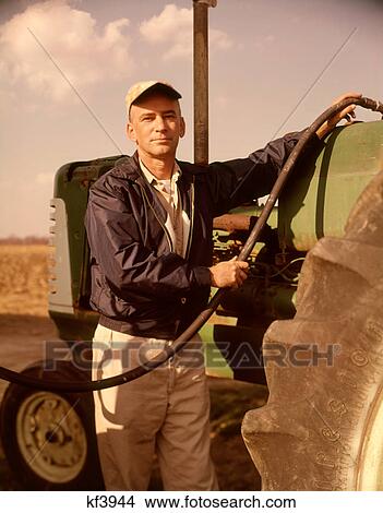 Picture - 1960s PORTRAIT OF SERIOUS MAN FARMER LOOKING AT CAMERA FUEL INTO TRACTOR. Fotosearch