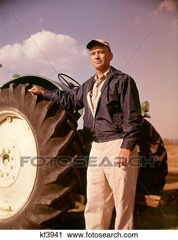 Stock Image - 1960s PORTRAIT OF SERIOUS MAN FARMER STANDING BESIDE LARGE TRACTOR TIRE LOOKING AT CAMERA. Fotosearch