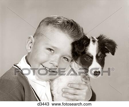 Stock Image - 1960s PORTRAIT OF SMILING BOY LOOKING AT CAMERA HUGGING HIS BLACK AND WHITE BEST FRIEND PET DOG HEAD TO HEAD. Fotosearch