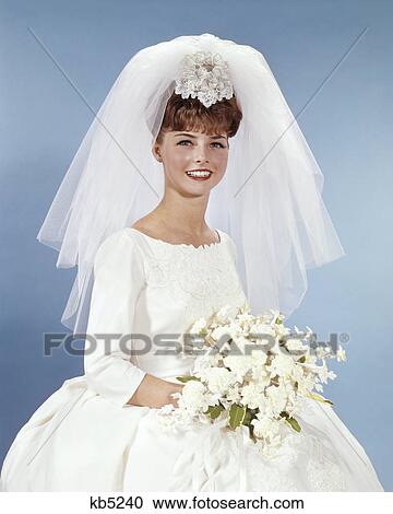 Stock Image - 1960s PORTRAIT SMILING WOMAN BRIDE IN SIMPLE WHITE WEDDING GOWN HOLDING WHITE FLOWER BOUQUET LOOKING AT CAMERA. Fotosearch
