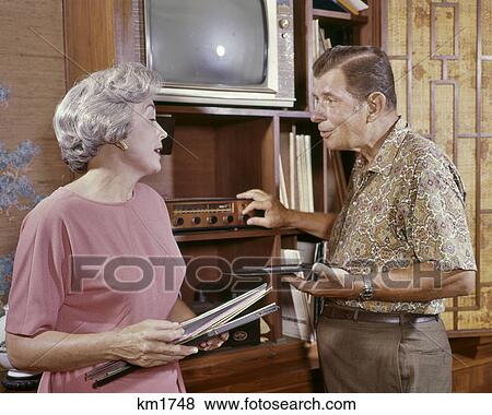 Stock Photo - 1960s SENIOR COUPLE LISTENING TO RECORDED MUSIC MAN TURNING STEREO PHONOGRAPH DIAL WOMAN HOLDING LP RECORD ALBUM COVERS. Fotosearch