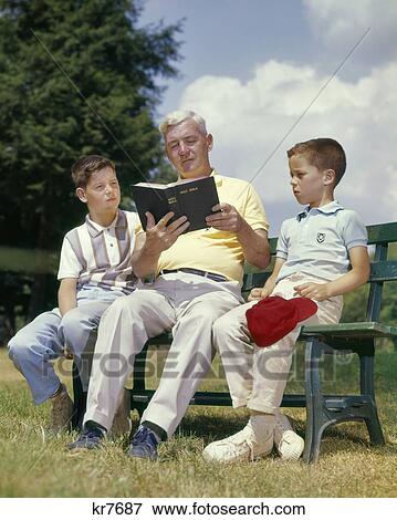 1960s SENIOR MAN GRANDFATHER READING BOOK BIBLE SITTING ON BENCH WITH TWO BOYS GRANDSONS View Large Photo Image Stock Photo - 1960s SENIOR MAN GRANDFATHER READING BOOK BIBLE SITTING ON BENCH WITH TWO BOYS GRANDSONS. Fotosearch