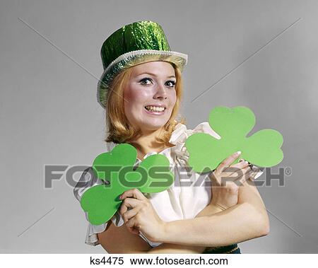 Stock Photography - 1960s SMILING BLOND WOMAN LOOKING AT CAMERA WEARING GREEN FOIL TOP HAT HOLDING TWO SAINT PATRICK DAY HOLIDAY IRISH SHAMROCKS. Fotosearch