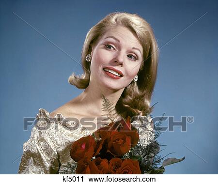 Stock Image - 1960s SMILING BLONDE WOMAN WEARING GOLD BROCADE BLOUSE DIAMOND EARRINGS HOLDING BOUQUET OF RED ROSES. Fotosearch
