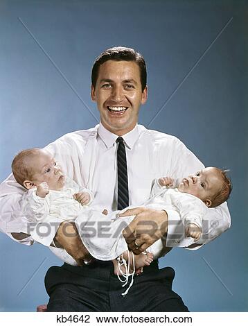 Stock Image - 1960s SMILING FATHER LOOKING AT CAMERA HOLDING TWO TWIN BABIES IN ARMS. Fotosearch