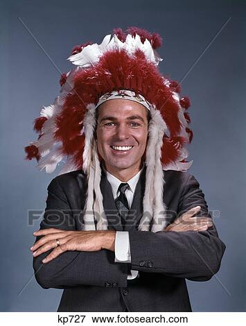 Stock Photo - 1960s SMILING MAN WEARING SUIT AND TIE AND NATIVE AMERICAN INDIAN FEATHER HEADDRESS ARMS FOLDED LOOKING AT CAMERA. Fotosearch