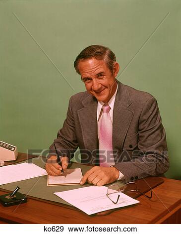 1960s SMILING MIDDLE AGED WHITE-COLLAR BUSINESSMAN SITTING WRITING AT OFFICE DESK GLANCING UP LOOKING AT CAMERA View Large Photo Image Stock Photo - 1960s SMILING MIDDLE AGED WHITE-COLLAR BUSINESSMAN SITTING WRITING AT OFFICE DESK GLANCING UP LOOKING AT CAMERA. Fotosearch