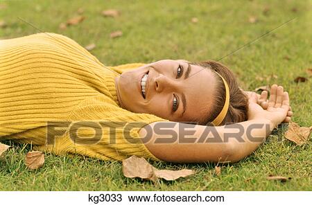 Stock Image - 1960s SMILING YOUNG BRUNETTE WOMAN LYING IN GRASS AUTUMN LEAVES LOOKING AT CAMERA WEARING YELLOW POOR BOY SWEATER AND HEADBAND. Fotosearch