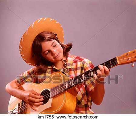Stock Photography - 1960s TEENAGER GIRL WEARING COWBOY HAT AND PLAID SHIRT PLAYING GUITAR. Fotosearch