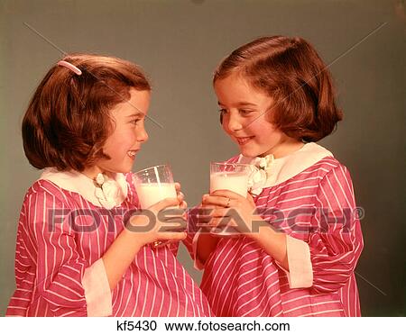Stock Image - 1960s TWIN SMILING BRUNETTE GIRLS WEARING PINK STRIPED DRESSES HOLDING DRINKING GLASSES MILK. Fotosearch