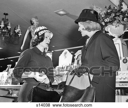 Stock Photo - 1960s TWO WOMEN WEARING HATS SHOPPING STANDING MEETING TALKING VISITING IN FRONT OF COSMETIC COUNTER OF DEPARTMENT STORE. Fotosearch