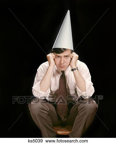 1960s UNHAPPY YOUNG MAN SITTING ON STOOL WEARING DUNCE CAP LOOKING AT CAMERA View Large Photo Image Stock Photo - 1960s UNHAPPY YOUNG MAN SITTING ON STOOL WEARING DUNCE CAP LOOKING AT CAMERA. Fotosearch