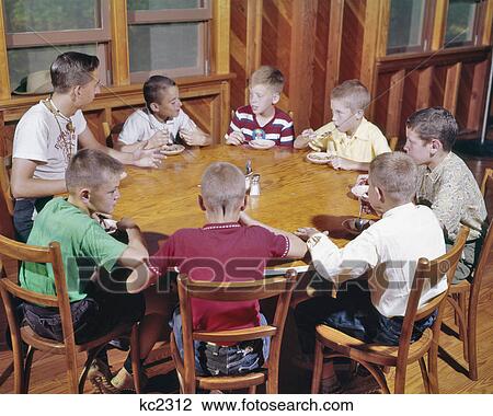 Stock Image - 1960s YOUNG TEENAGE MAN CAMP COUNCILOR SITTING AT WOODEN ROUND TABLE EATING ICE CREAM TALKING WITH SEVEN KIDS BOY CAMPERS. Fotosearch