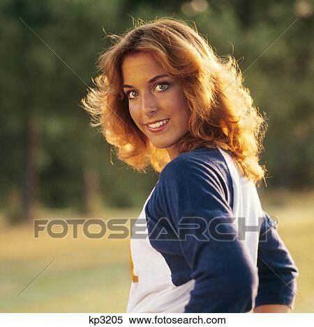Stock Photography - 1970s 1980s SMILING YOUNG WOMAN SUNLIGHT ON HER GOLDEN HAIR LOOKING AT CAMERA OVER HER SHOULDER. Fotosearch