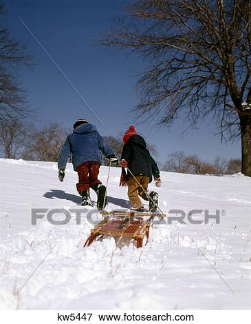 Stock Photo - 1970s 2 TWO BOYS PULLING WOODEN SLED UP HILL WINTER SNOW KIDS. Fotosearch